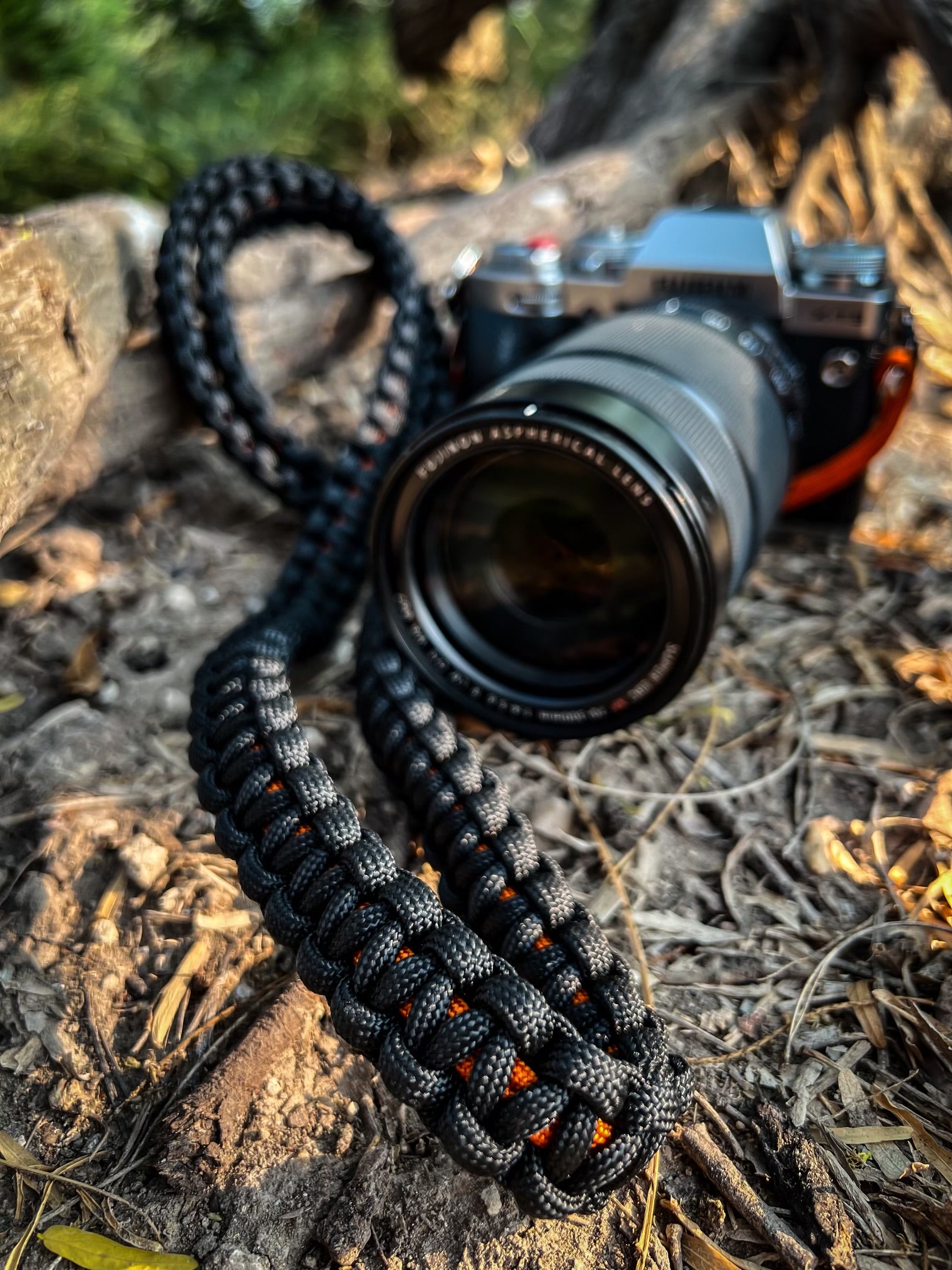 Photo of the orange and black "Altamira Oriole" Venture Strap used as a camera strap. The strap and camera are on a log in the outdoors. The Venture Strap is a binocular or camera strap made of sturdy paracord. The strap is worn across the body and is more comfortable and durable that traditional binocular harnesses or camera straps.