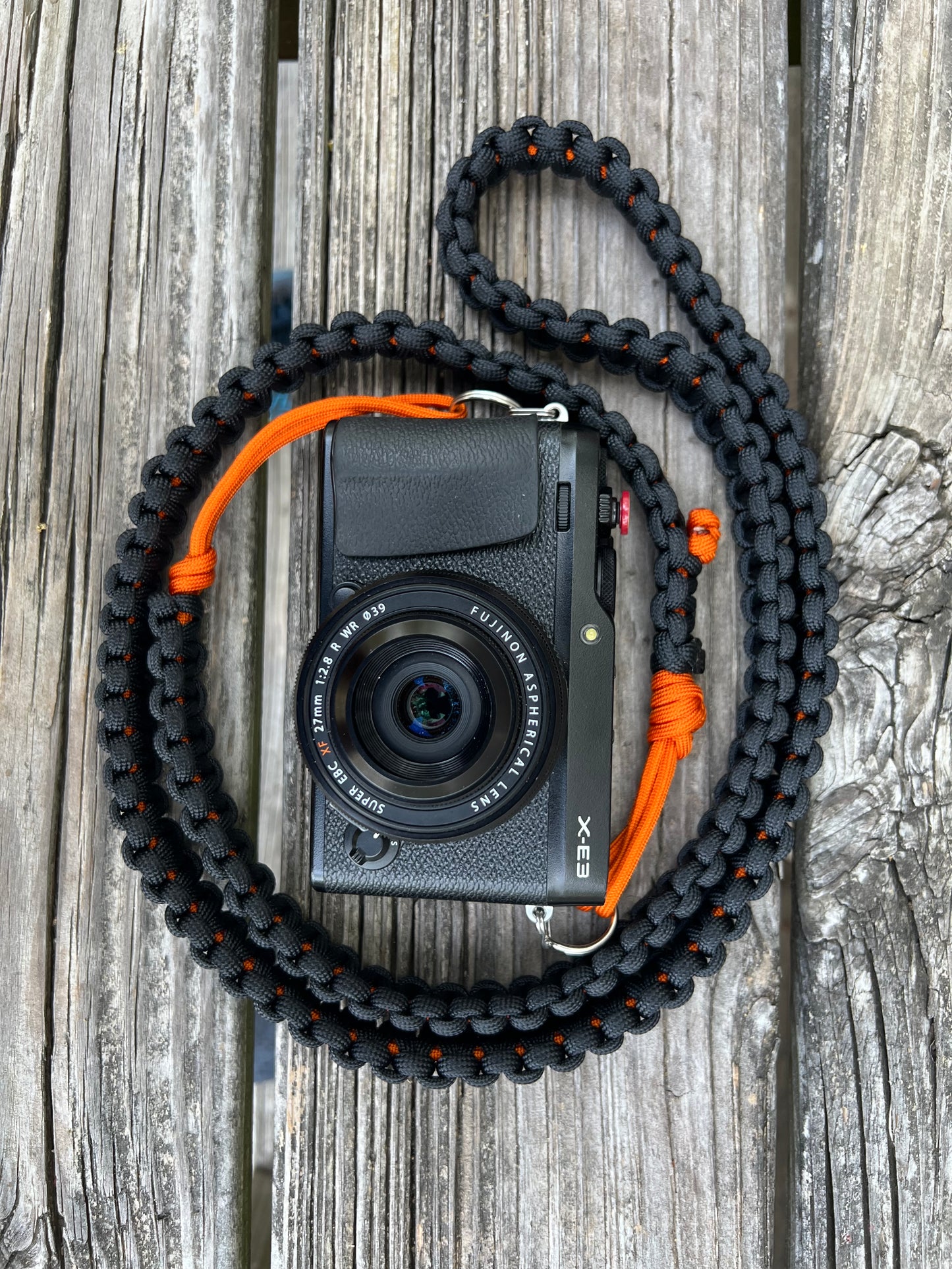 An orange and black "Atlamira Oriole" Venture Strap attached to a viewfinder style camera. The strap and camera are artfully posed on a picni bench in the outdoors. The Venture Strap is a binocular or camera strap made of sturdy paracord. The strap is worn across the body and is more comfortable and durable that traditional binocular harnesses or camera straps.