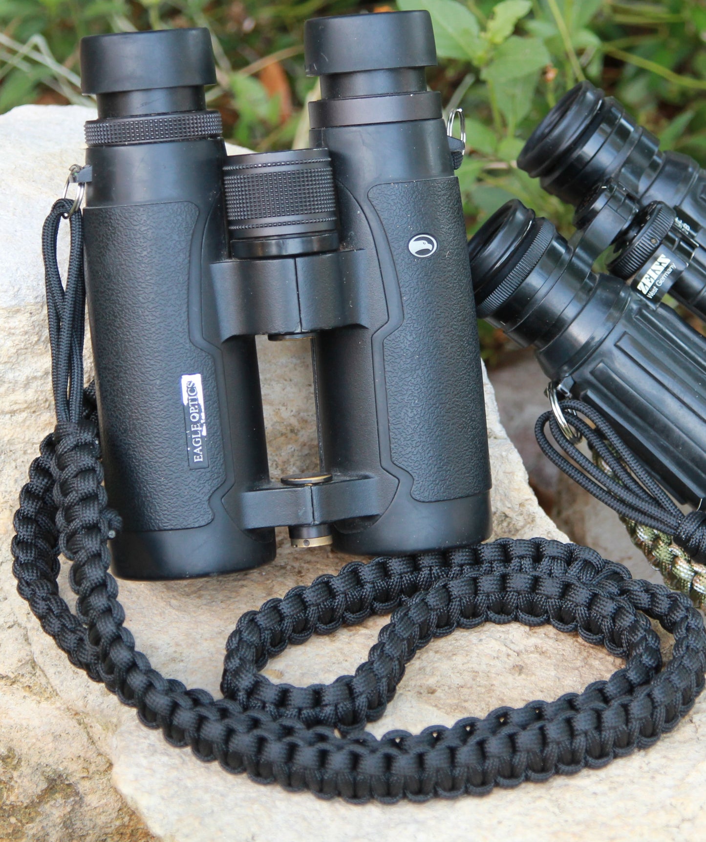 The Venture Strap is a binocular or camera strap made of sturdy paracord. The strap is worn across the body and is more comfortable and durable that traditional binocular harnesses or camera straps. This particular photo features the all black "Groove-billed Ani" color. The strap is attached to a pair of binoculars leaning on a rock.