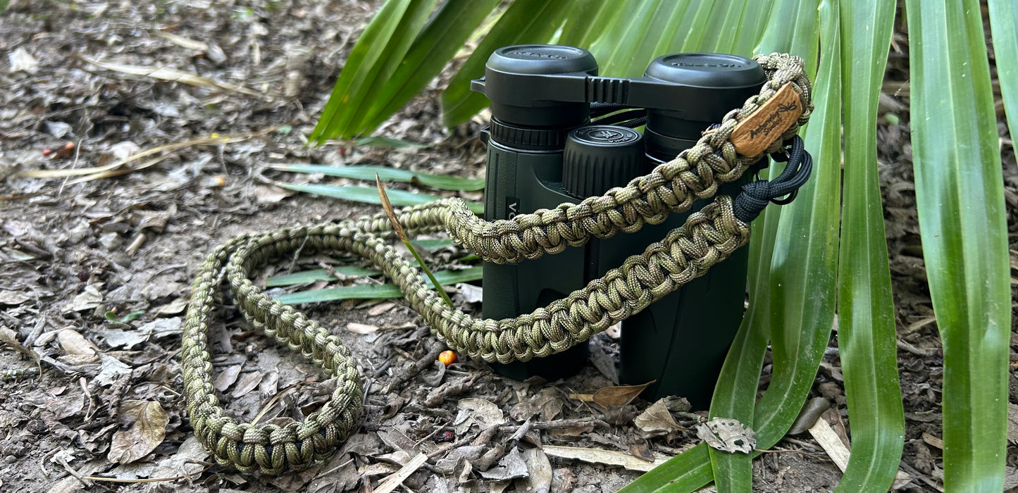 The Venture Strap is a binocular or camera strap made of sturdy paracord. The strap is worn across the body and is more comfortable and durable that traditional binocular harnesses or camera straps. This photo shows The Venture Strap in the Olive Sparrow color that features classic camo greens and browns. The strap is connected to a pair of binoculars which are set on the ground next to a palm frond. 